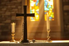 Cómo usar esta Oración Milagrosa para la Salud Mental, muestra una cruz de madera con un rosario transparente, acompañada de dos velas encendidas, frente a vitrales coloridos en una iglesia.