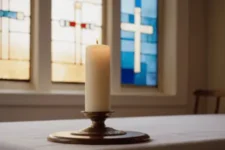 Oración al Padre Pío, muestra una vela encendida sobre una mesa blanca con un fondo de vitrales que destacan cruces azules y doradas, creando un ambiente sereno y espiritual.