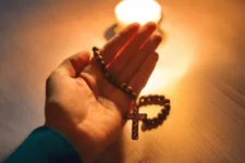 Oración de Sanación Física, muestra una mano sosteniendo un rosario con una cruz, iluminada por la luz cálida de una vela sobre una superficie de madera.