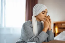 Oraciones para Pedir la Salud de un Enfermo, muestra a una mujer con un pañuelo en la cabeza, con las manos juntas en actitud de oración, sentada junto a una mesa en un ambiente cálido y tranquilo.