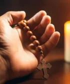 Padre Pio Oración Poderosa para Pedir Salud y Sanación Física, muestra una mano sosteniendo un rosario con una cruz metálica, iluminada por la luz cálida de una vela encendida al fondo.