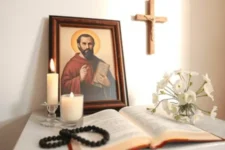 Quién es San Charbel y por qué se le invoca para la salud: Muestra una mesa con un cuadro de San Charbel, una cruz, flores blancas, velas encendidas y una Biblia abierta con un rosario negro.