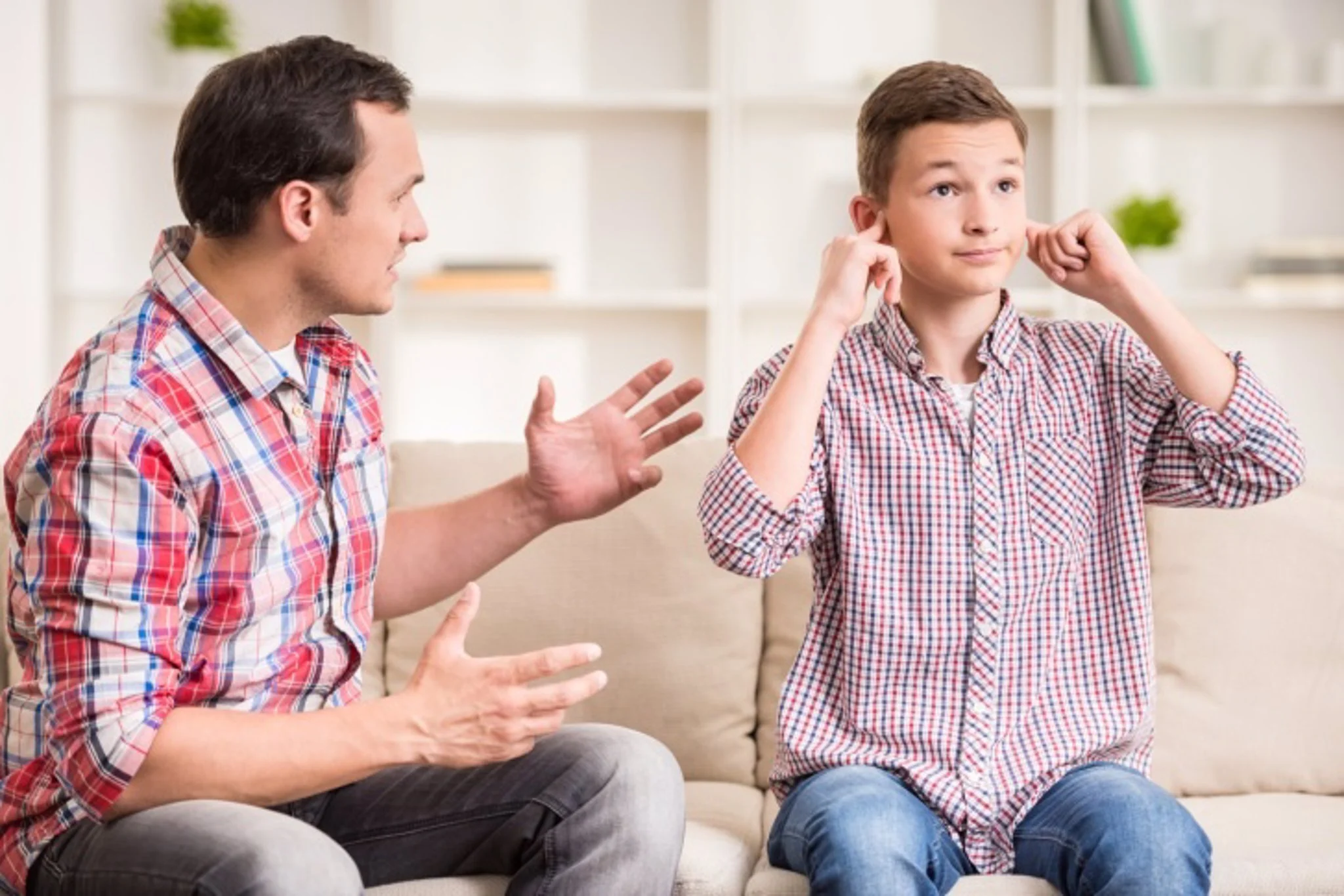 Un padre sentado en un sofá habla gesticulando a su hijo adolescente, quien bloquea sus oídos con los dedos, mostrando una actitud de resistencia, en una sala de estar con estanterías al fondo.